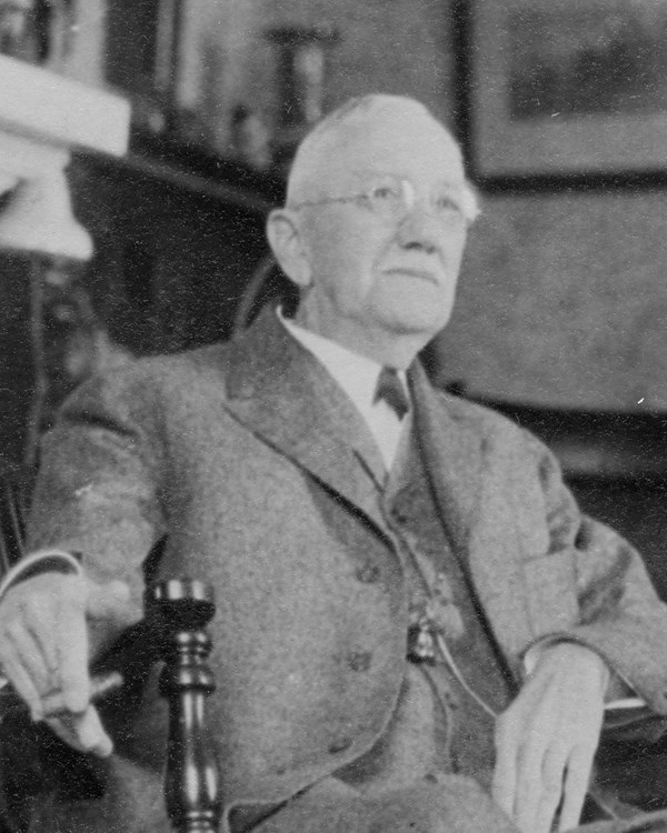 Black and white image of an older man wearing a suit sitting in a wooden chair.