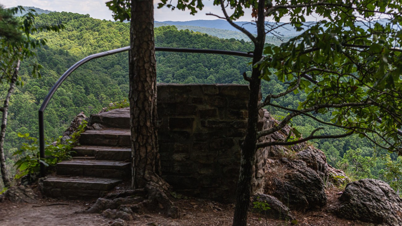 A black metal rail surrounds an outcropping overlooking rolling, forested hills.