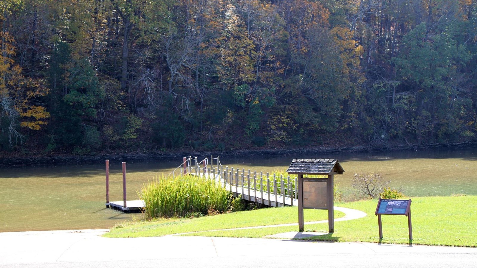 A dock sits in some water, a ramp to the left to deposit boats, 2 informational signs near road
