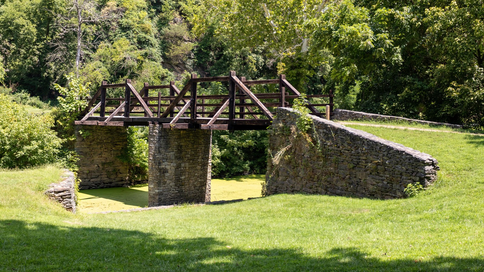 A wooden trestle on stone pillars crosses a creek into some woods.
