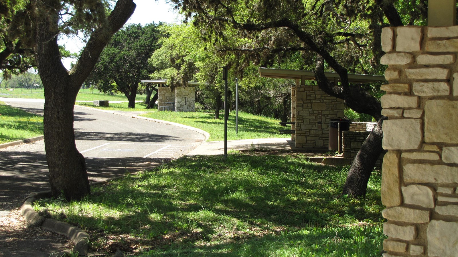 Off a curved driveway are three covered picnic shelters with stone walls under many shade trees.