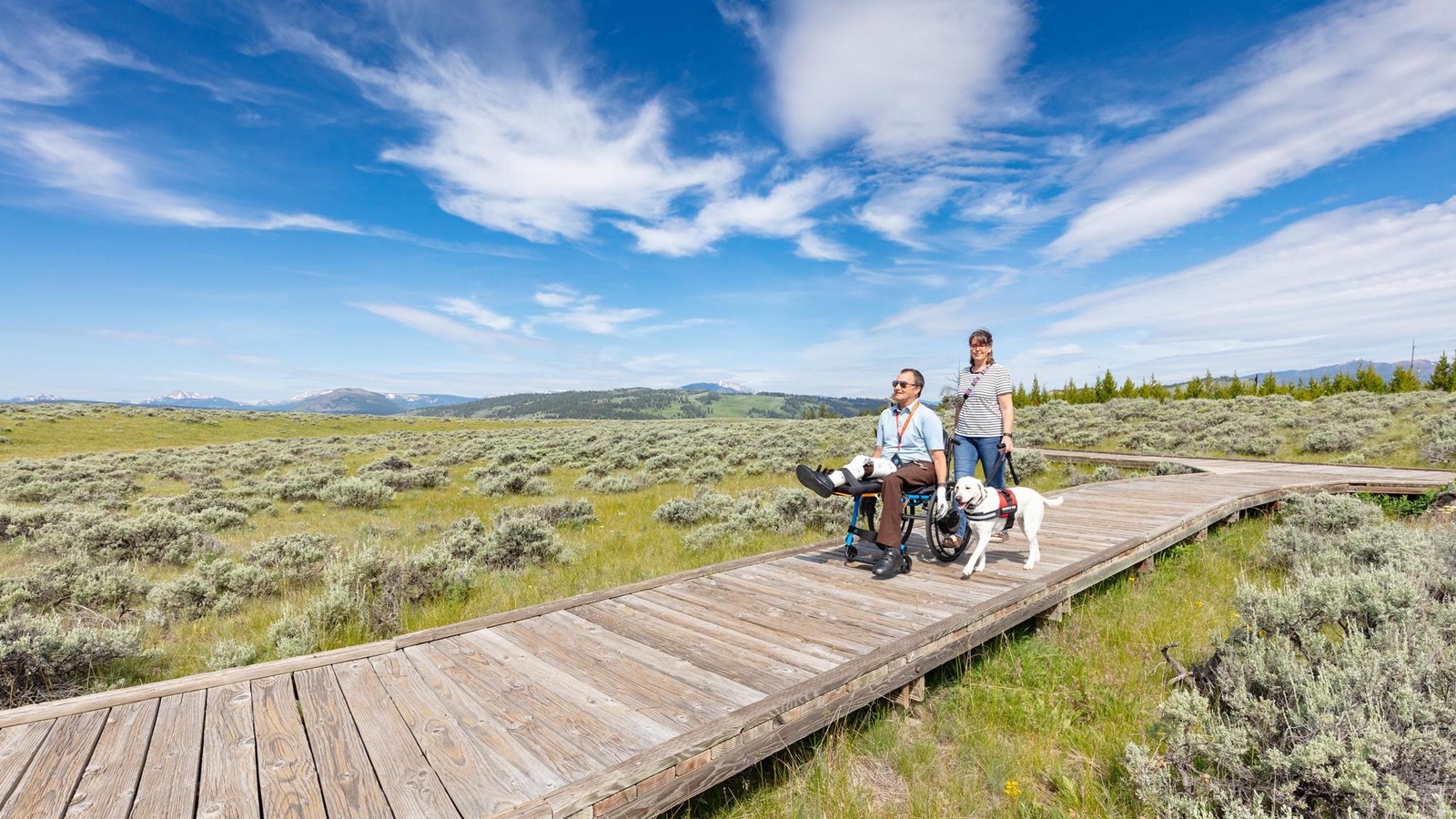 Two people, one in a wheelchair, and a service animal explore a boardwalk through the sagebrush.