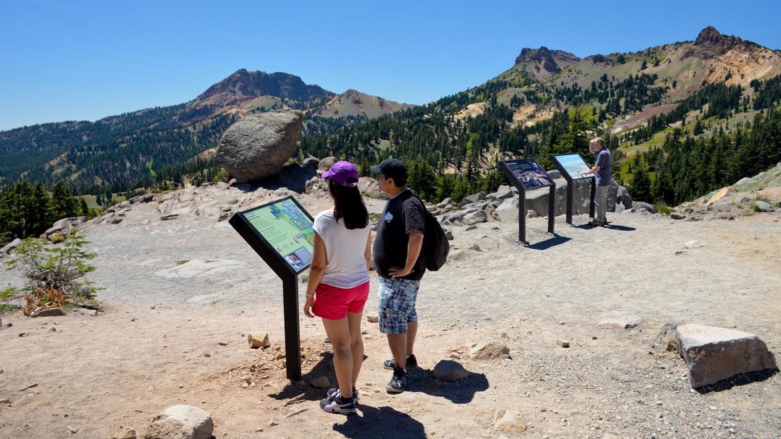 Visitors standing reading waysides about geology in the Bumpass Hell parking area with the glacial e