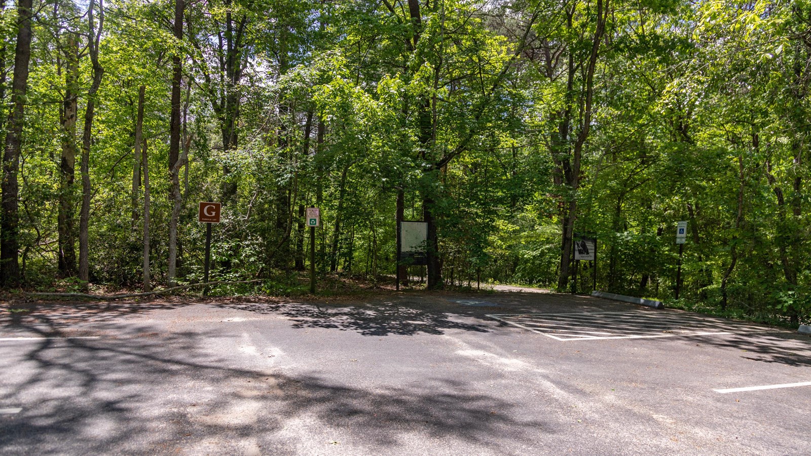 A brown sign with the letter G stands in front of a wayside and a paved parking lot in the woods
