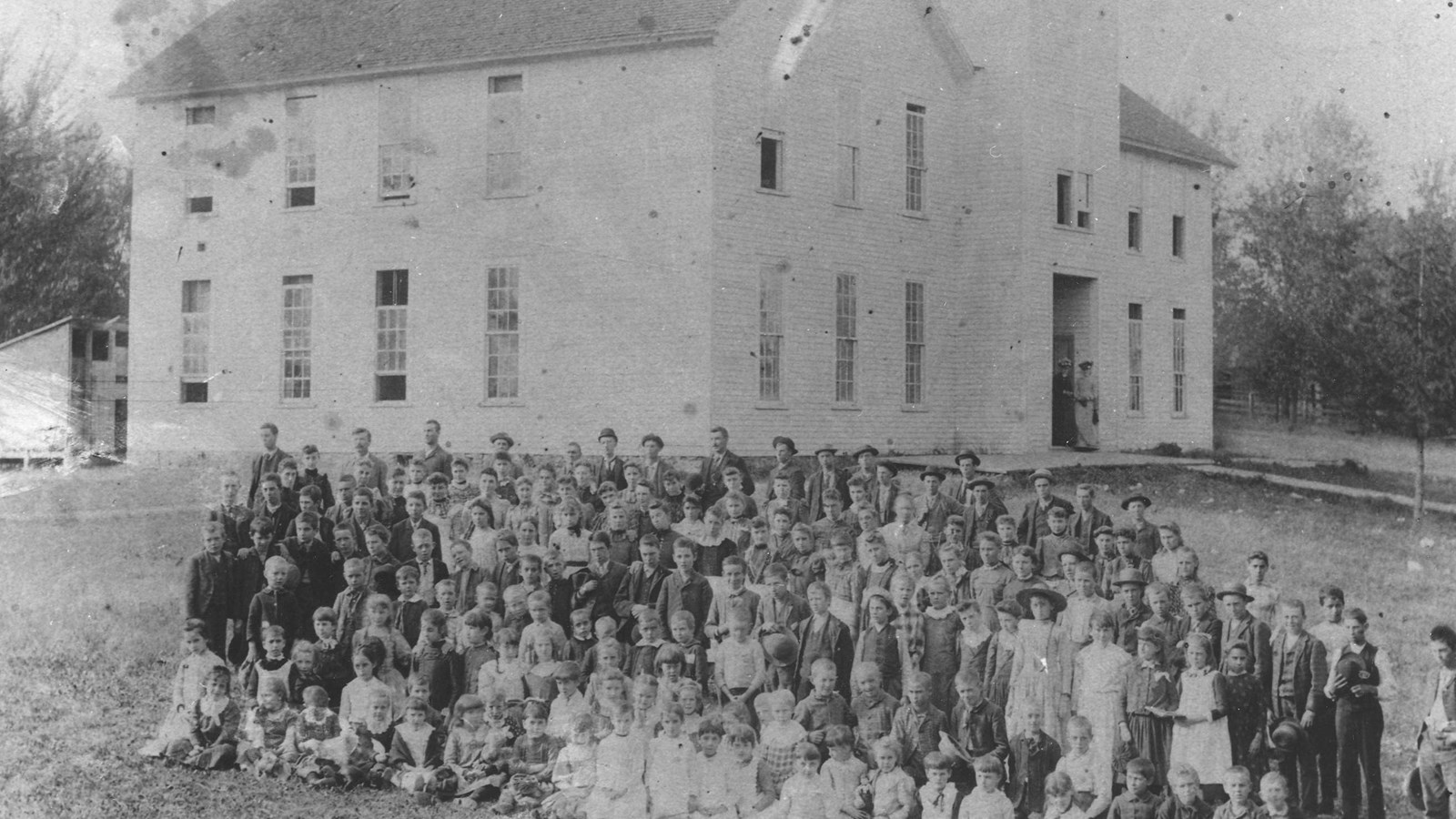 A black and white photo depicting a large two-story schoolhouse with students in the foreground.