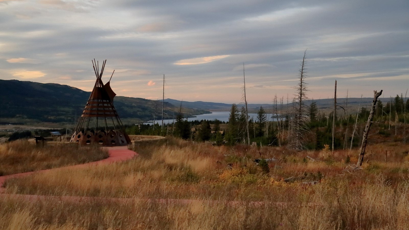 Photo of a grassy area at sunset with mountains and river available in the distance with metal tipi
