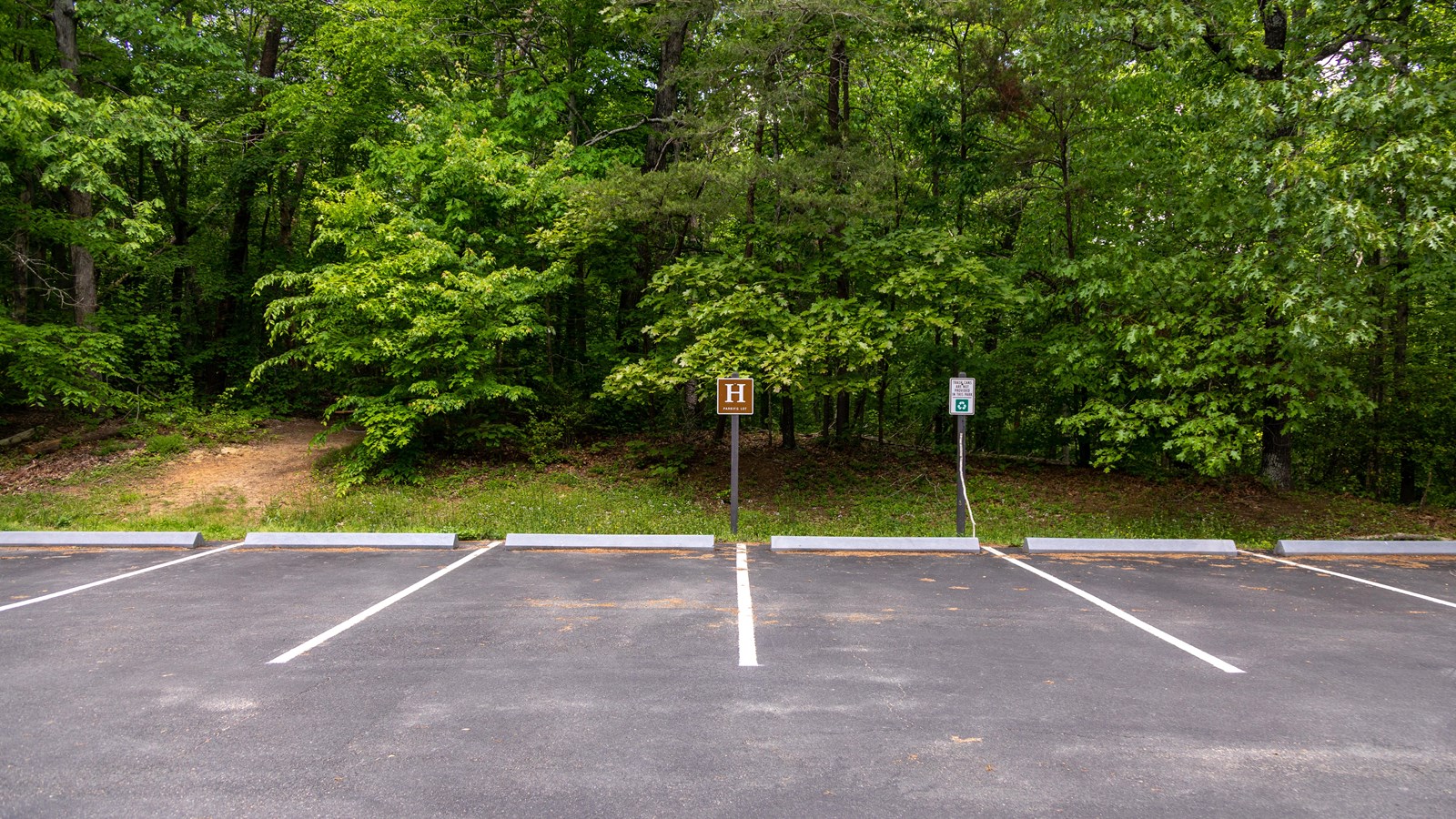 A brown sign with an H on it stands in front of a paved parking lot in the woods