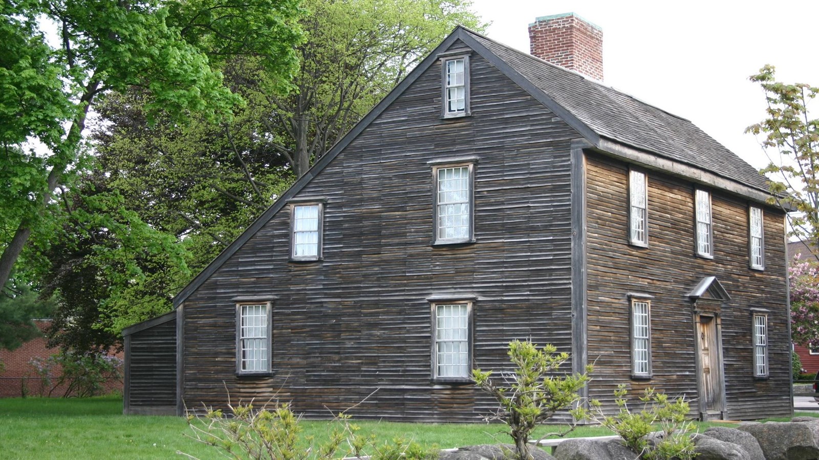 A brown saltbox style home with many windows and a chimney in the middle of the roof.