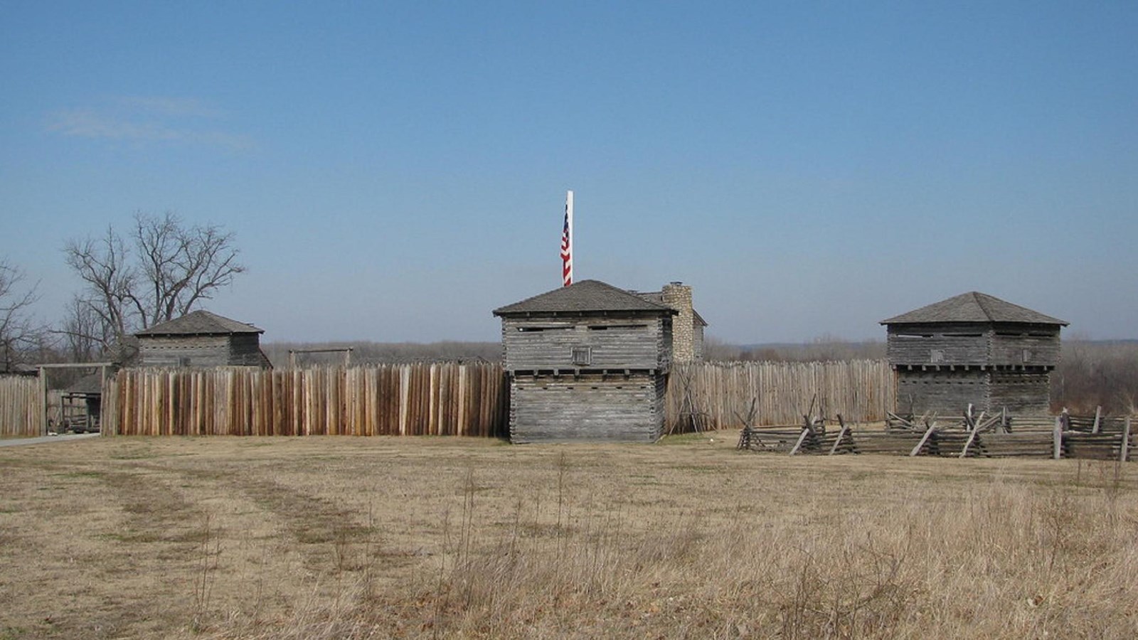 A natural wood palisade with square sentry towers surrounds a few old building