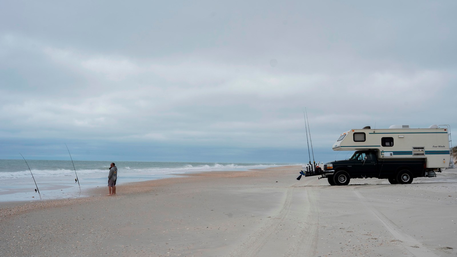 A camper truck and a person are on the beach. Cloudy skies behind them.