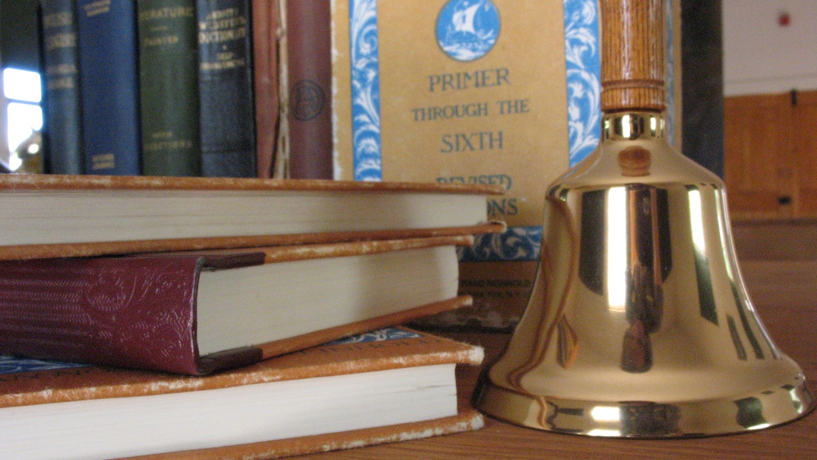 A shiny hand bell stands on a desk next to a stack of 1900s schoolbooks.