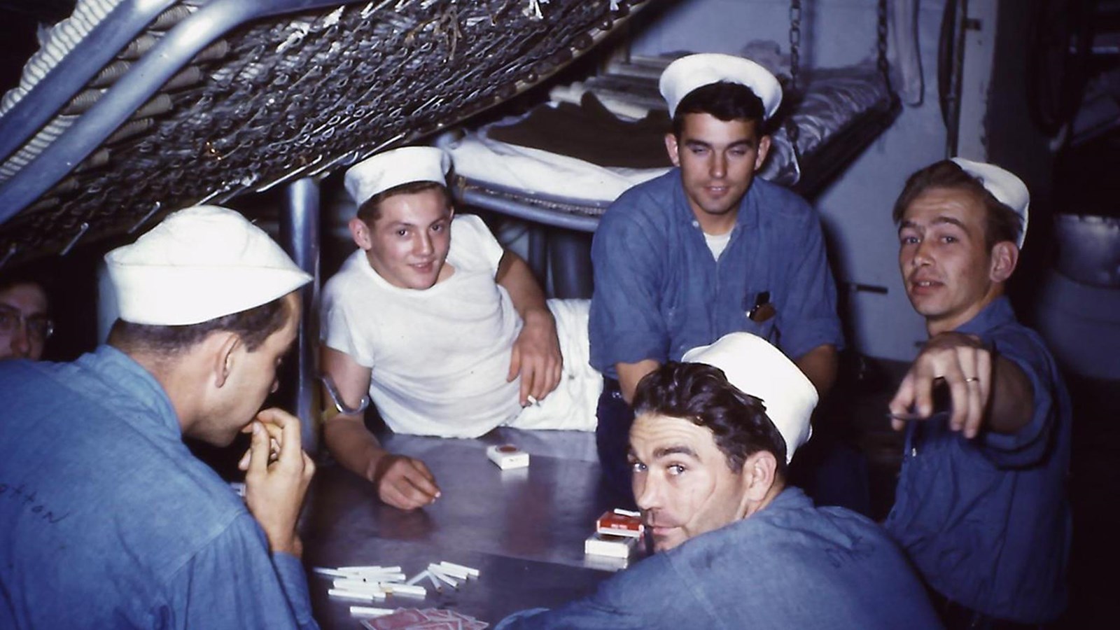 Five sailors in dungaree shirts, one in a white tee, with hats, sit around a card game