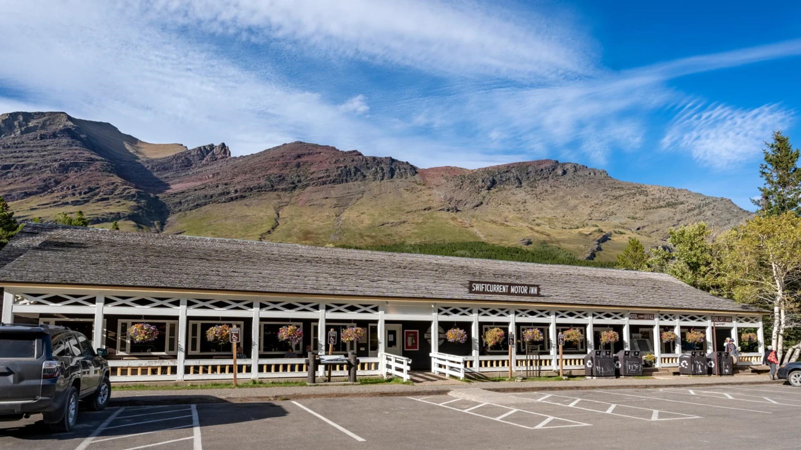 A rustic one-story motel with a white wooden porch sits in fron tof a mountain range.