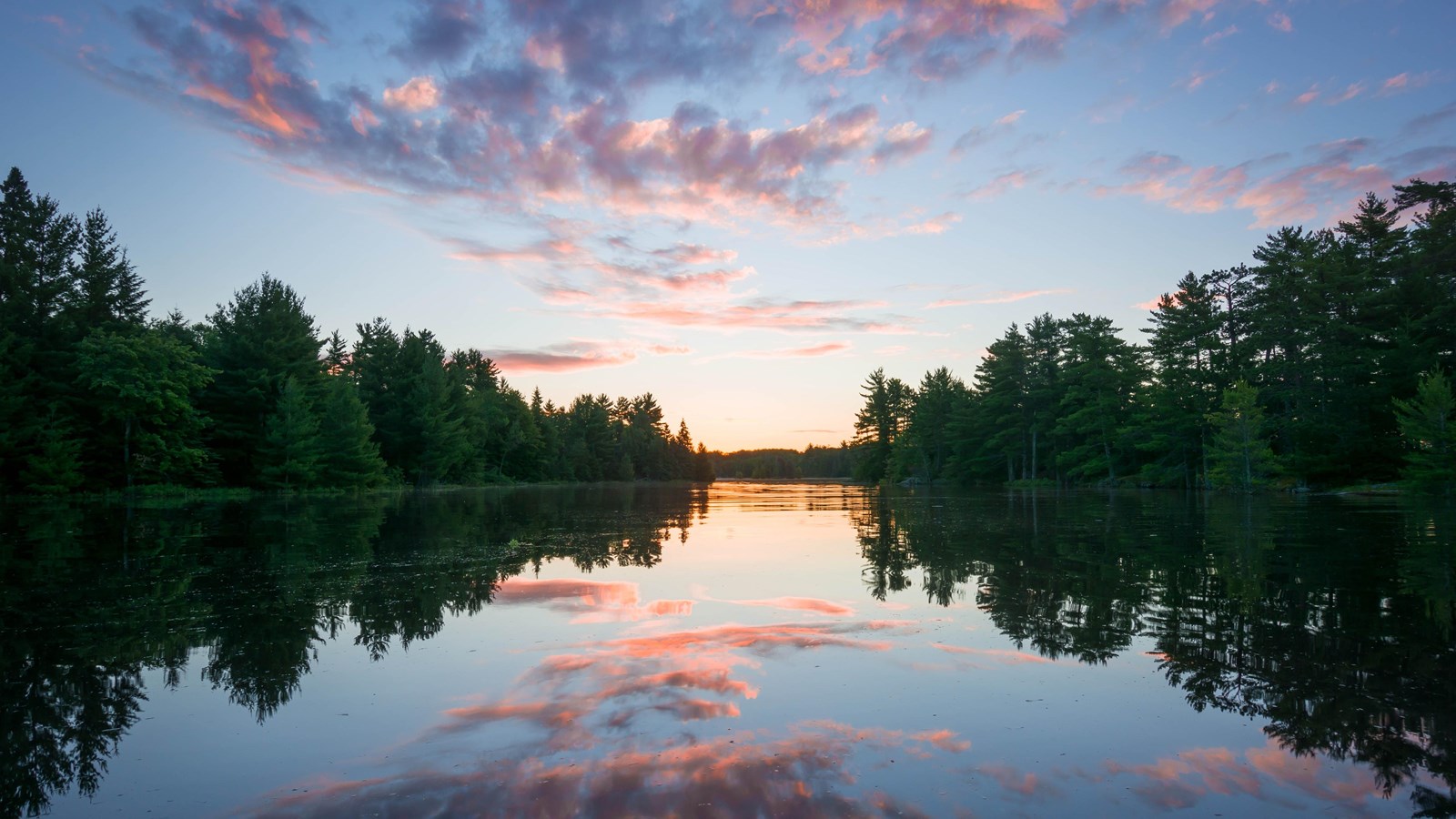 Clouds are reflected in a calm lake. Dense forest surrounds both sides of the lake.