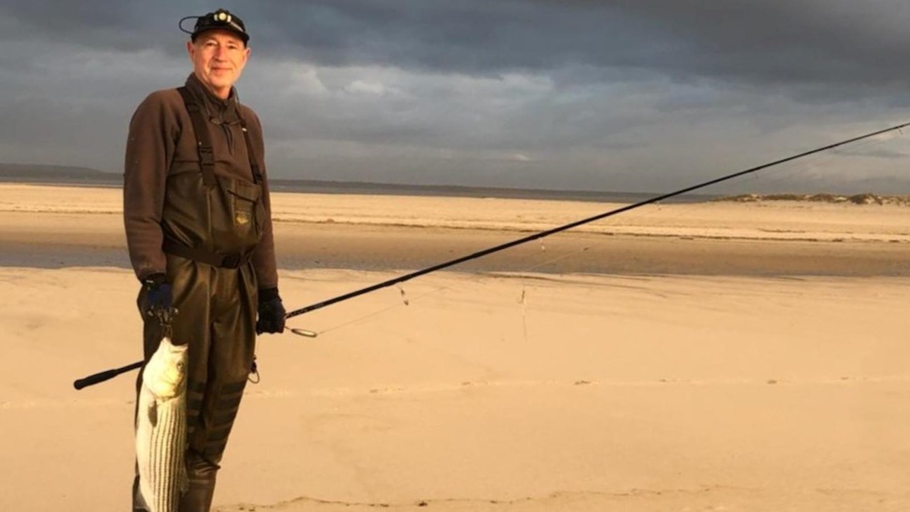 A fisherman stands smiling on an ocean beach with a large stripped bass he caught.