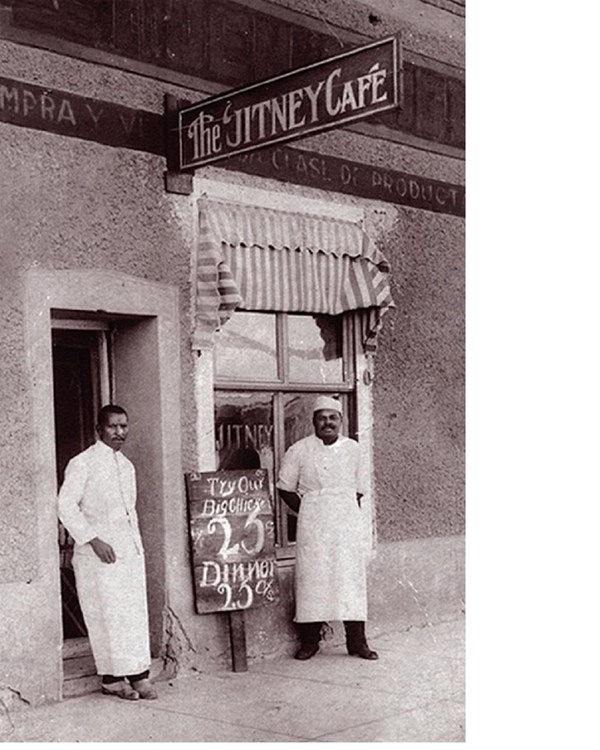 two men in long aprons stand outside of their café building with an advertisement sign