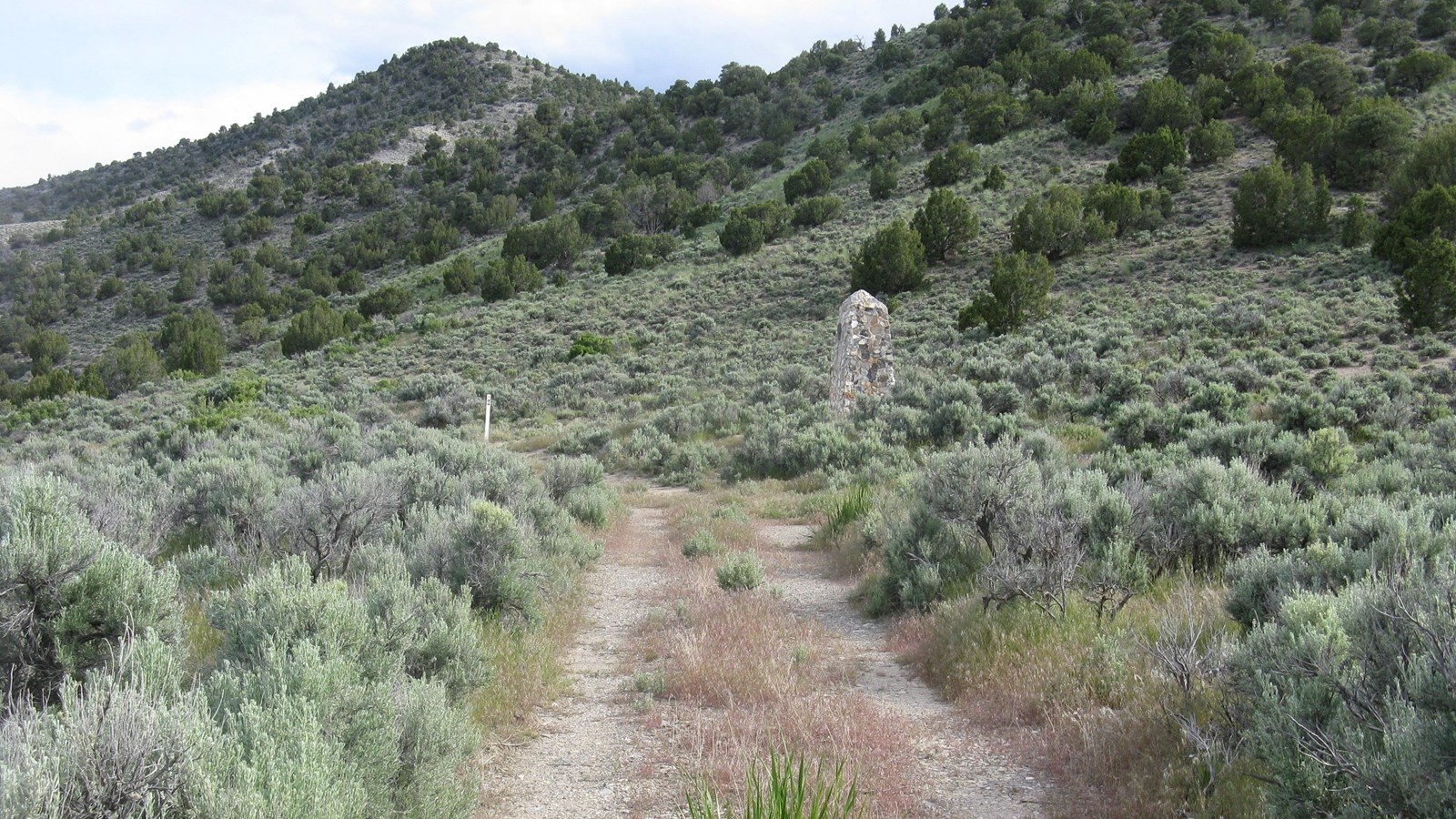 A stone monument rests along a sagebrush trail beneath rising slopes under an overcast sky.