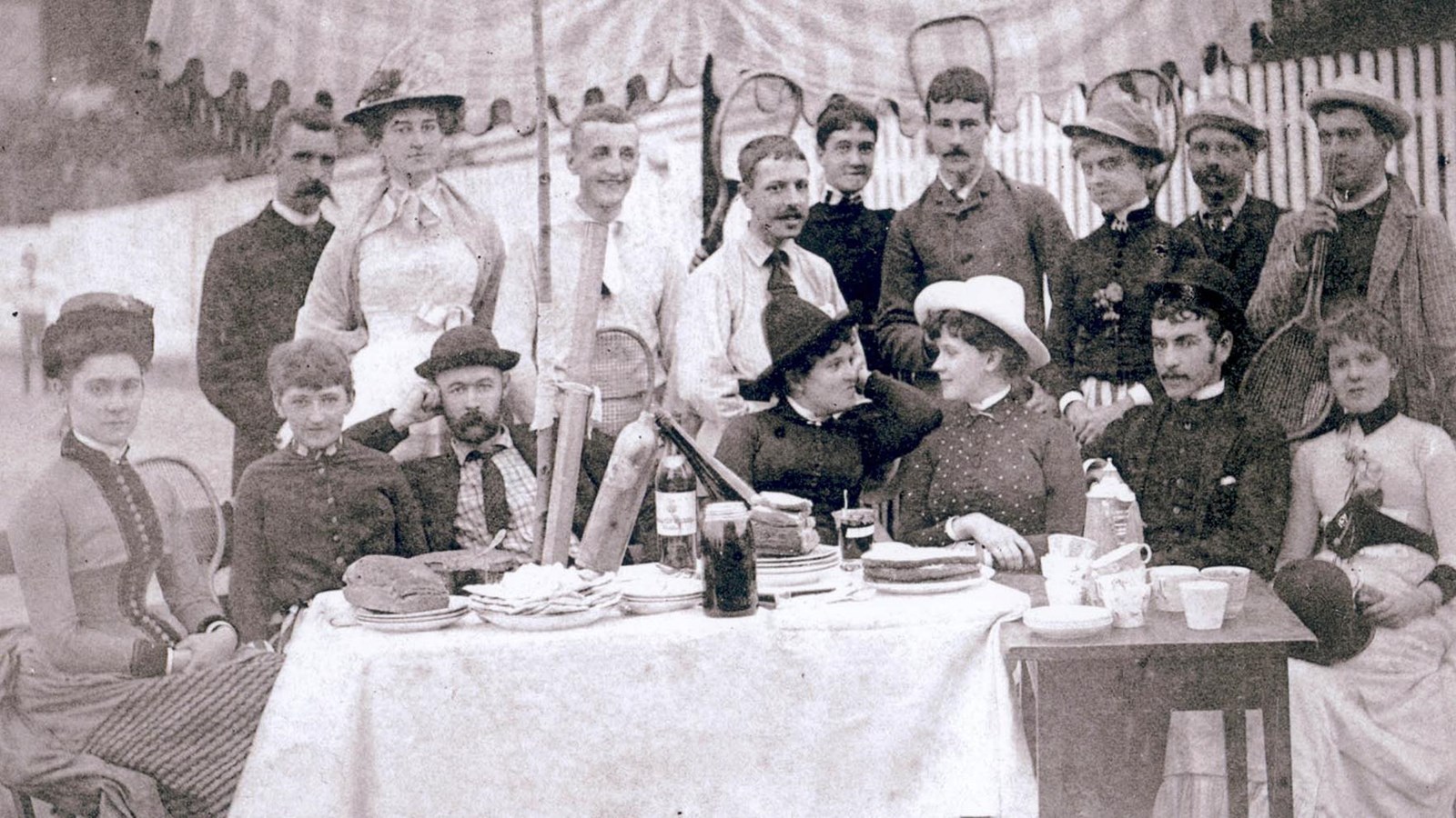 Men and women in 1920s fashion sit around a picnic table holding an assortment of dishes.