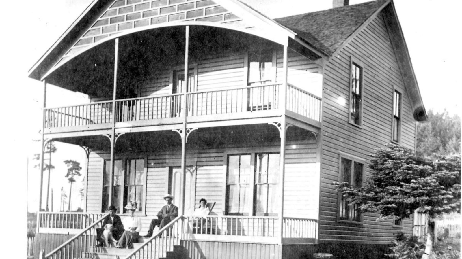 Black and white photograph of a large home constructed of wood and brick with people in front.