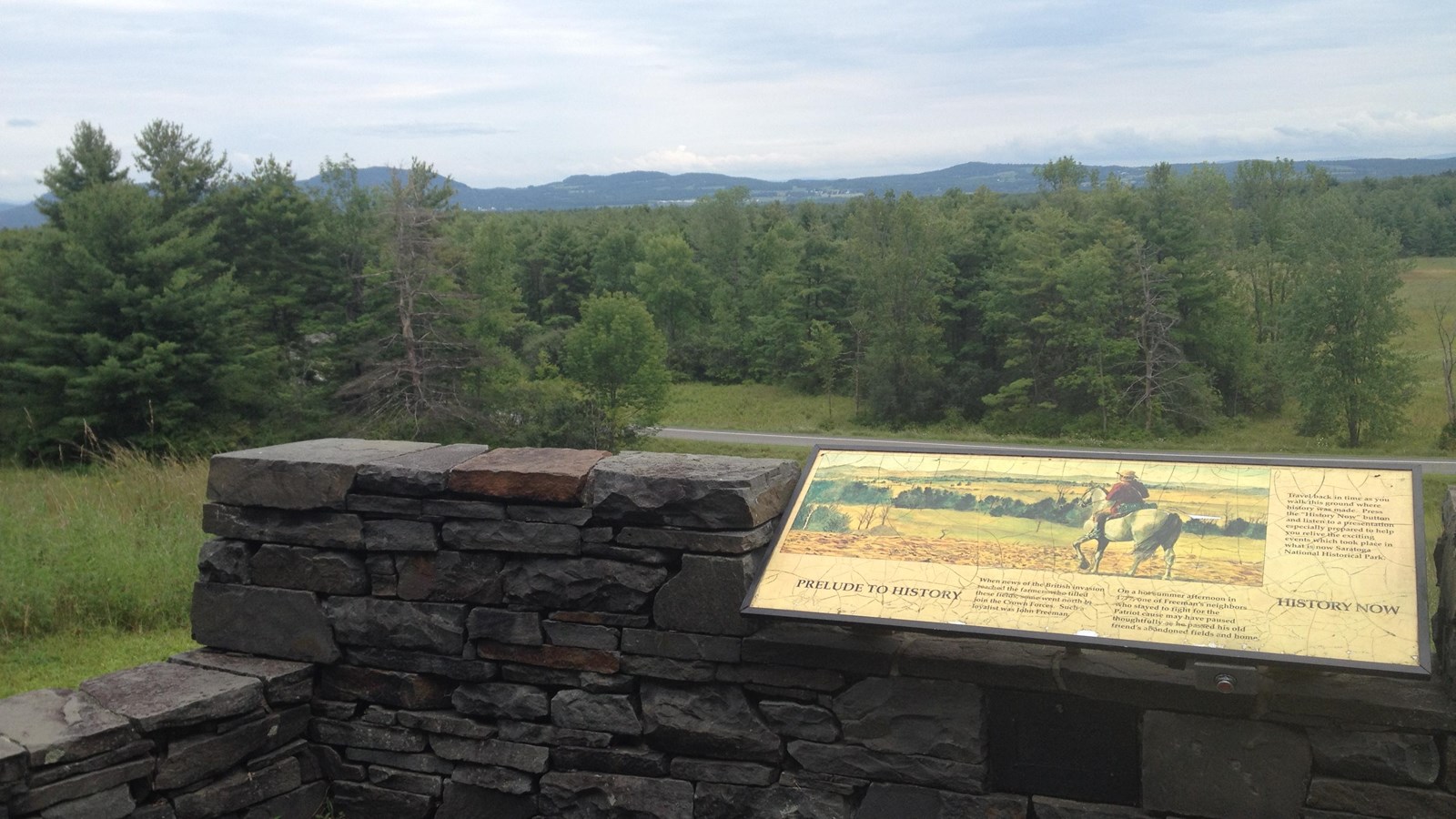 A stone wall with an informational sign overlooking a field covered in trees