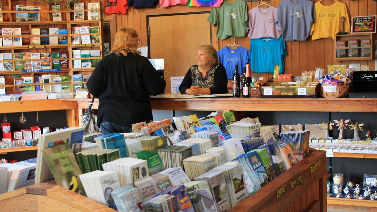 woman speaking to another woman behind a counter in a room filled with shirts, fliers, and souvenirs