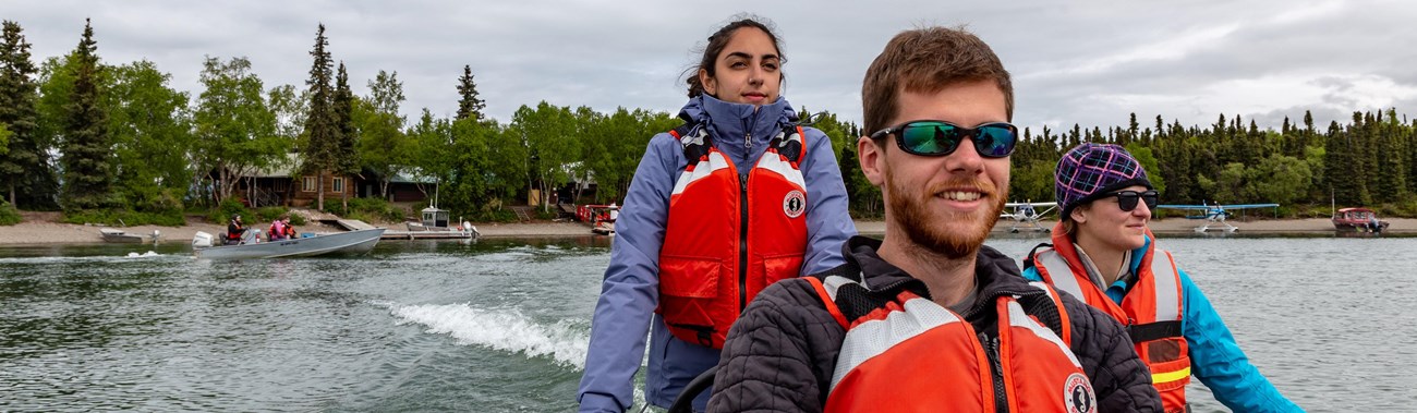 3 people wearing PFDs ride in a boat from the shore of Port Alsworth