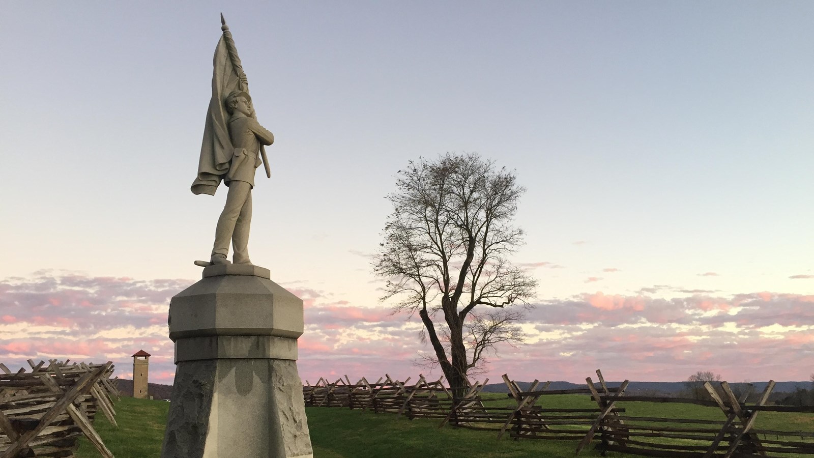 A stone statue with a man holding a flag stands next to a depression surrounded by a wooden fence.