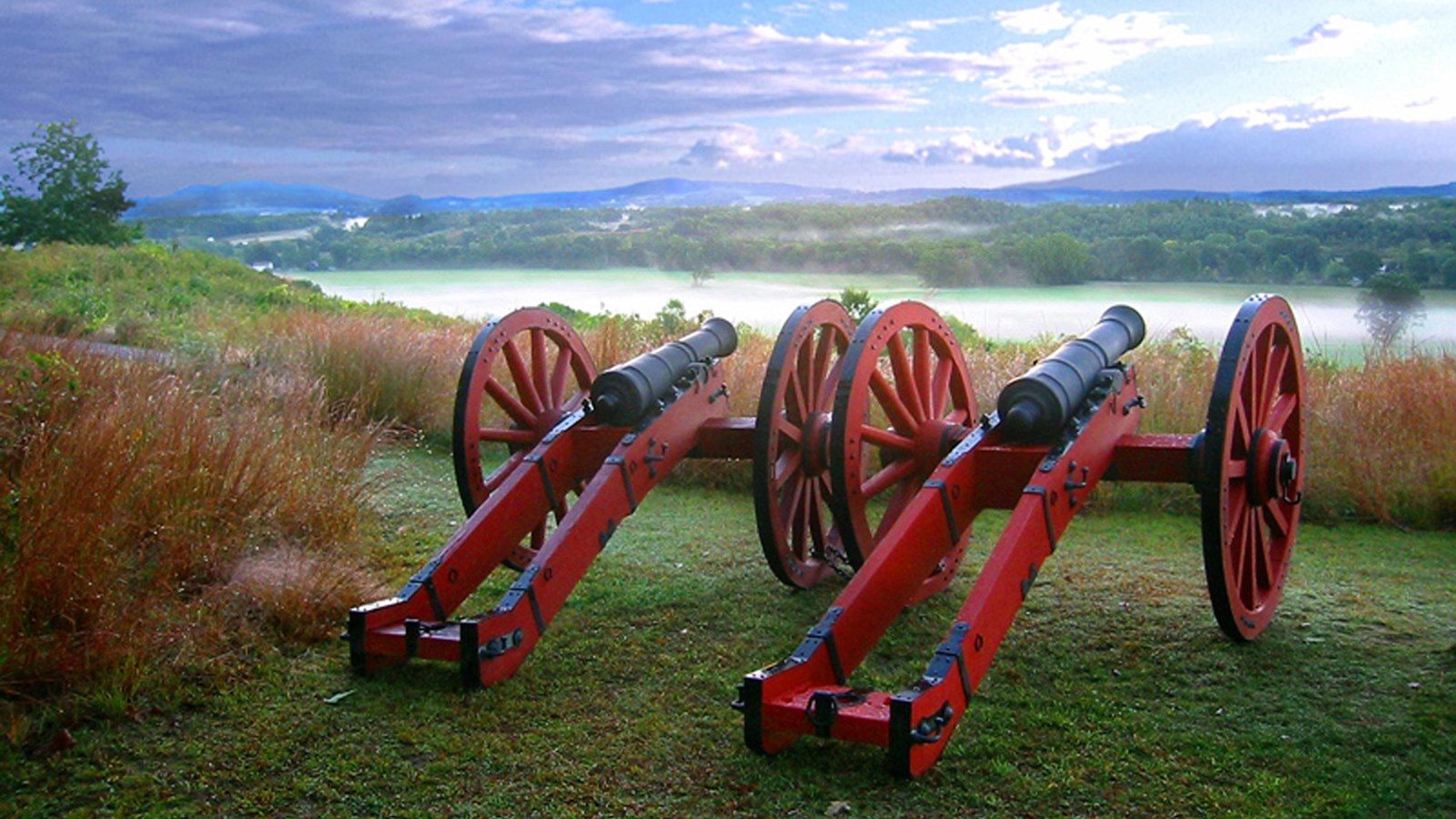 Two bright red cannons overlooking a fog covered field.
