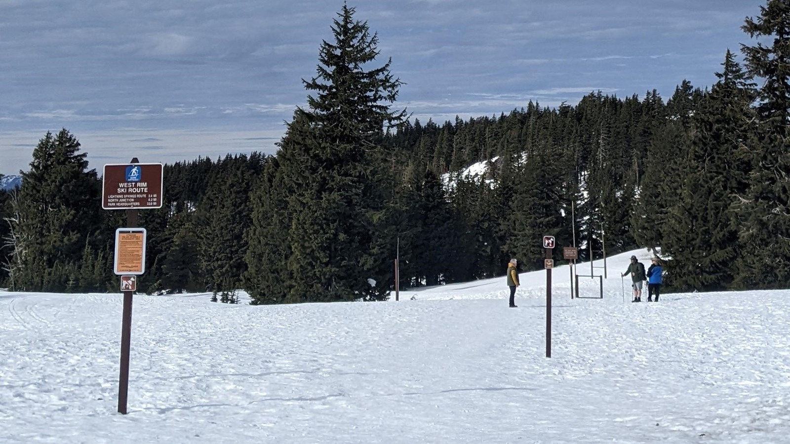 Snow covered road and landscape, a trail route information post in the snow people and trees in back