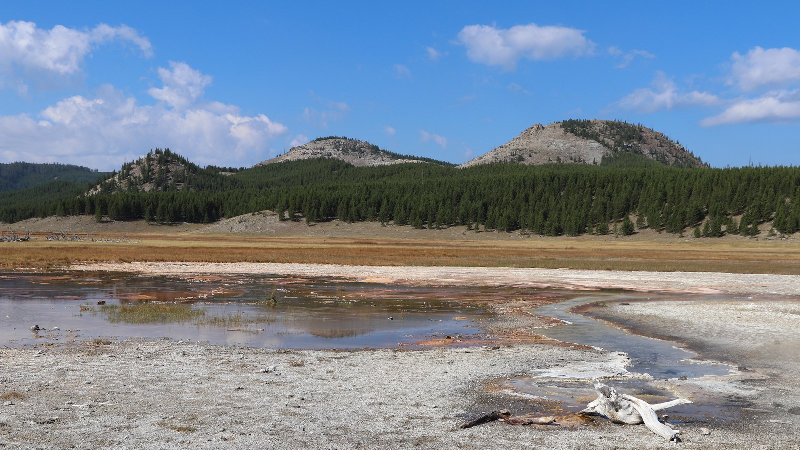 A thermal area sits in a meadow in front of rolling hills.
