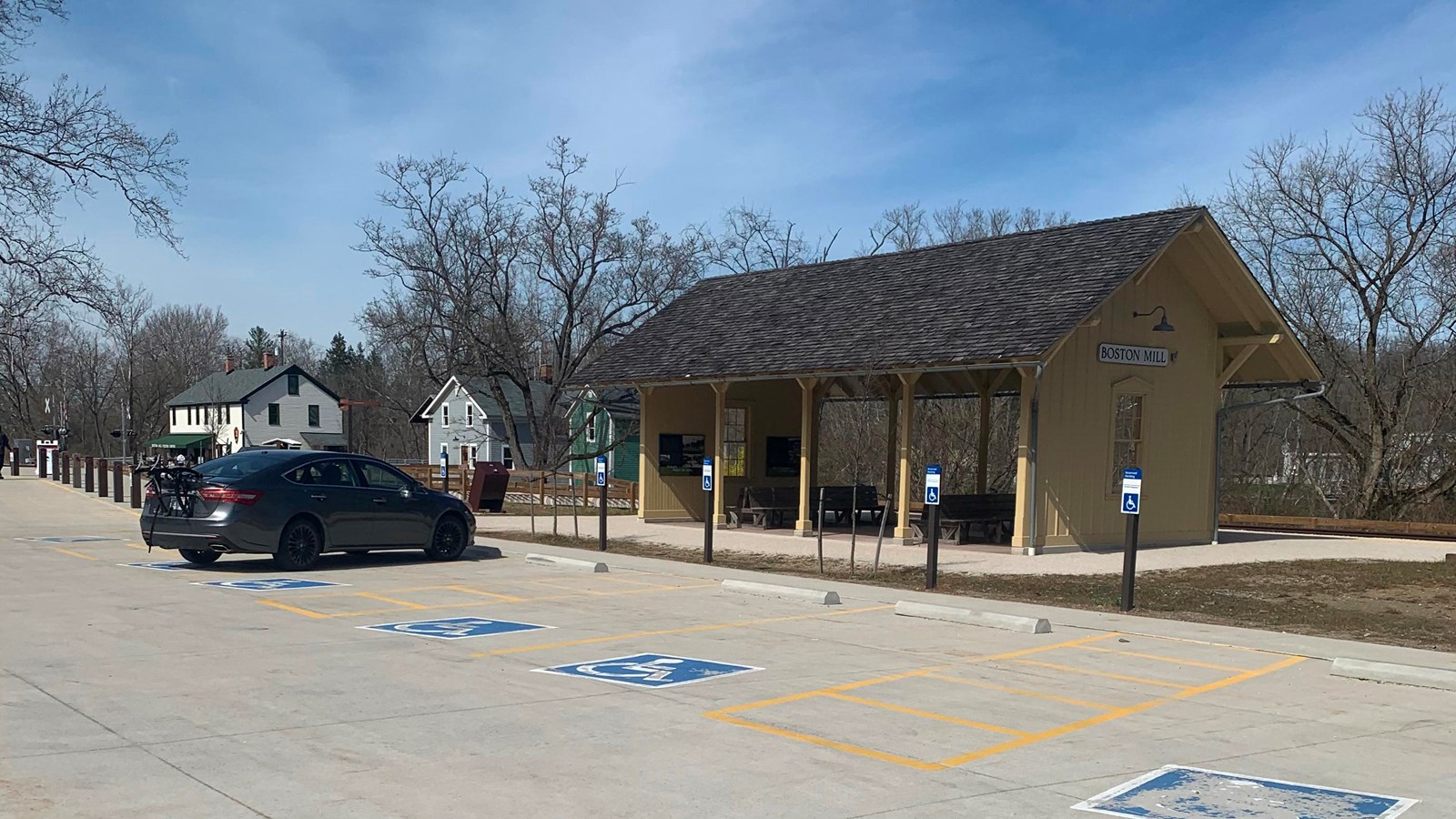 Yellow train station, open on two sides, with accessible parking in front and two buildings behind.