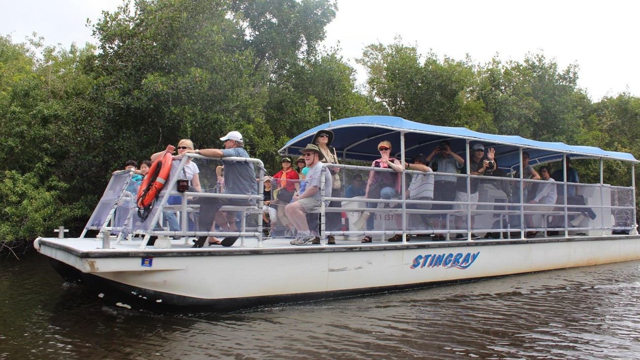 A large open air white boat with blue canopy top with passengers seated motors through murky water