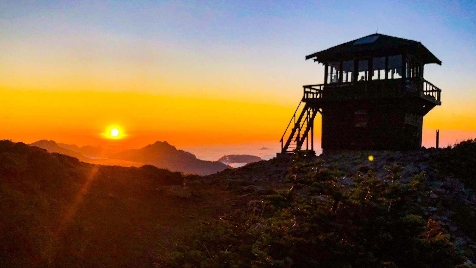 Silhouette of wooden fire lookout against colorful sunset with mountains