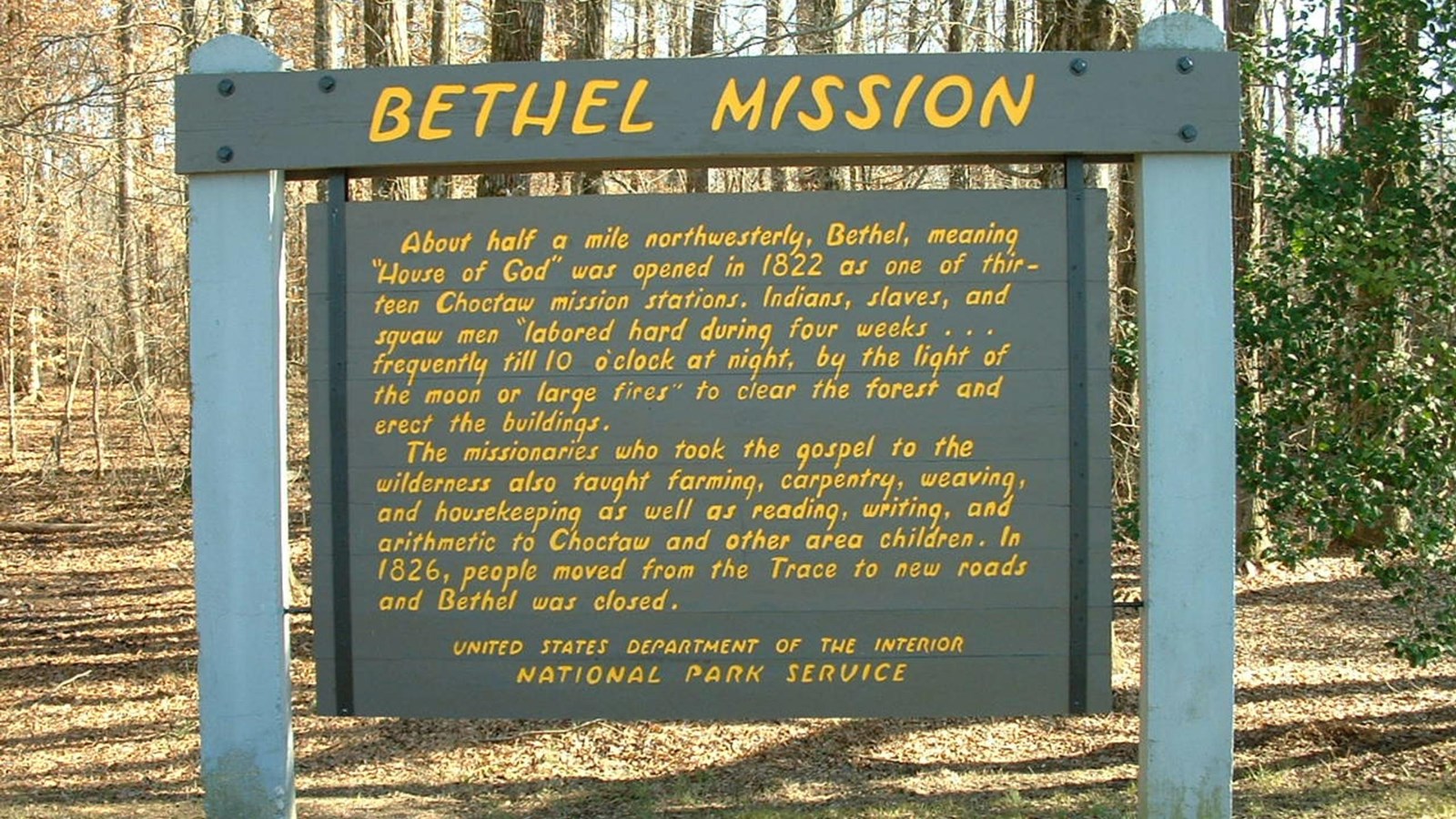 A brown sign with yellow text on concrete posts  in front of  a forest. 
