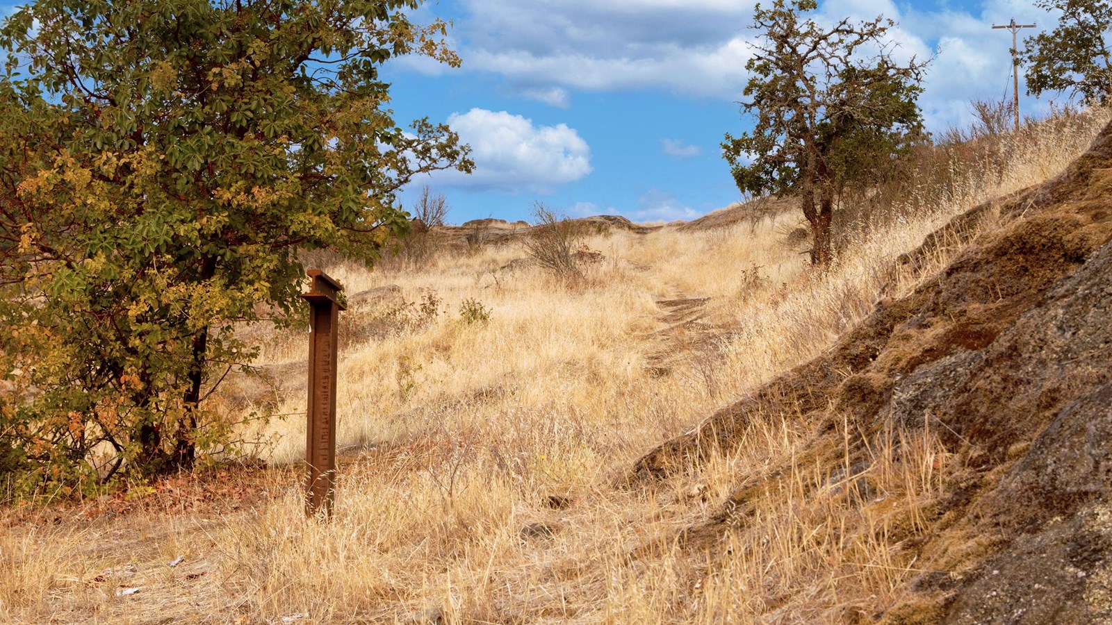 Trail follow a rut carved into the rock on a hill slope. Short yellow grass covers the slope with a 