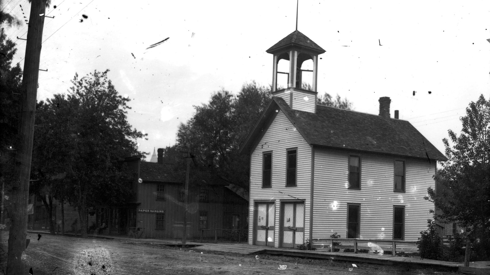 A false-front building stands across the street from a white firehouse with a bell tower.