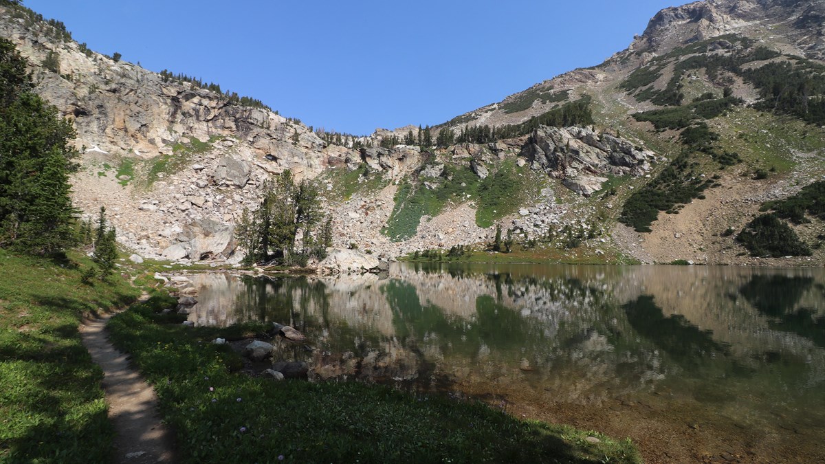 An alpine lake sits at the base of a rocky cliff surrounded by green vegetation.