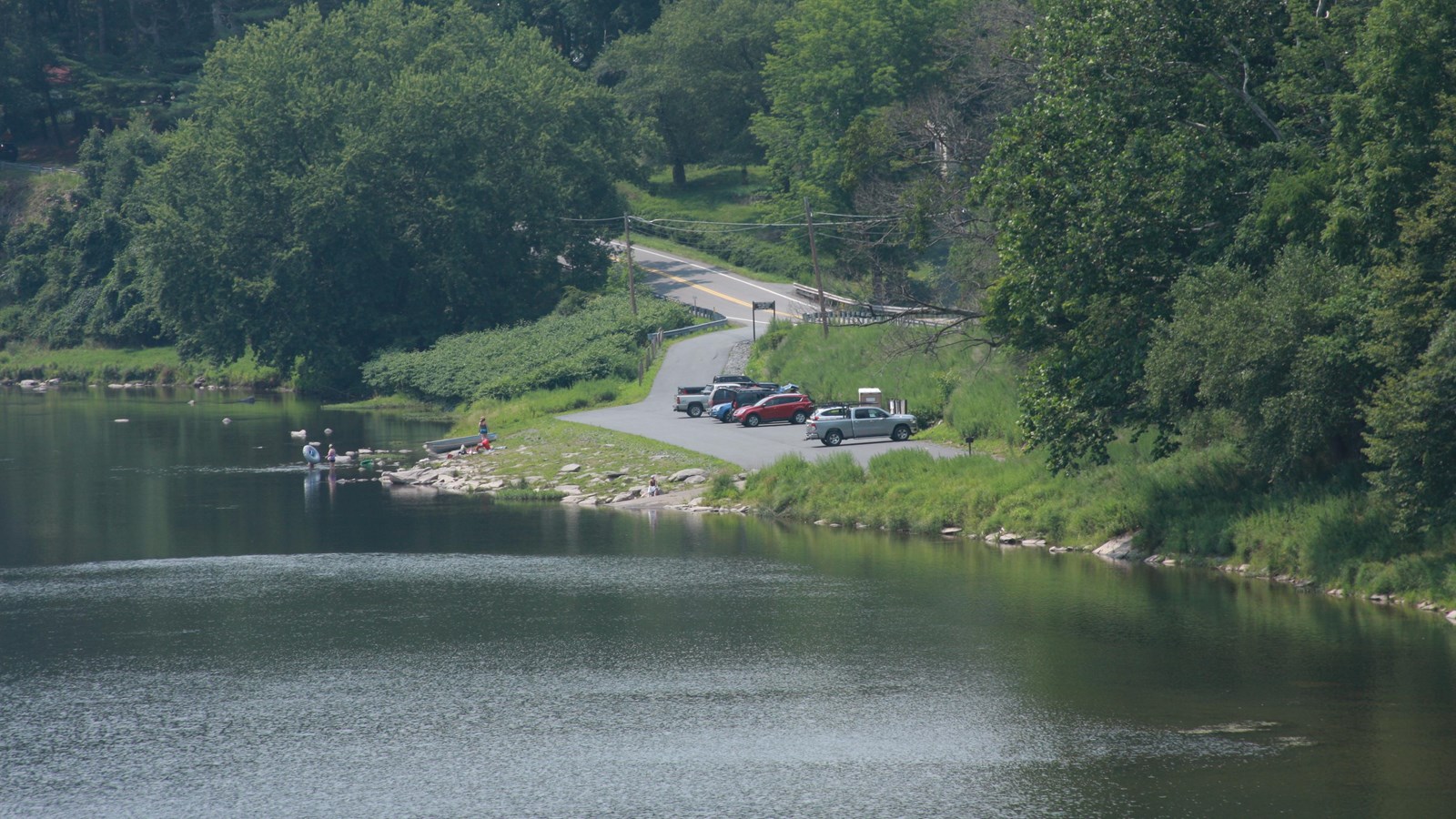 View of riverbank with paved area parallel to water with cars. Several people with tubes and a boat.