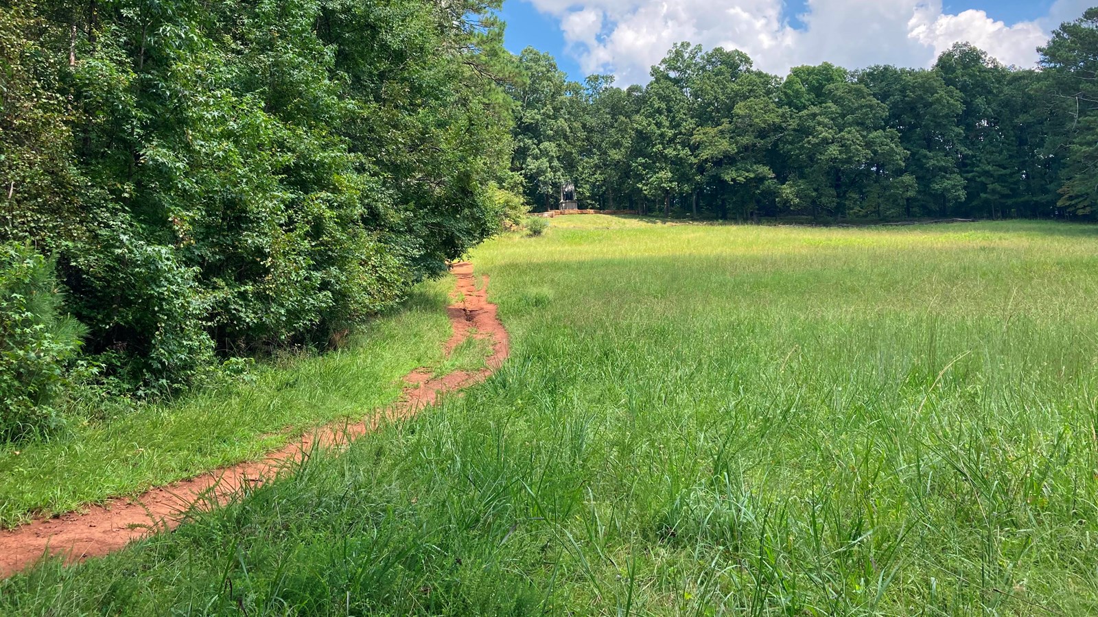 A reddish orange dirt path at the edge of a large green field snakes up a steep hill.