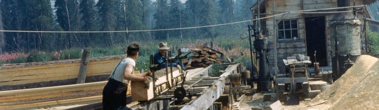 Two men working on sawmill stacked lumber in foreground and background.