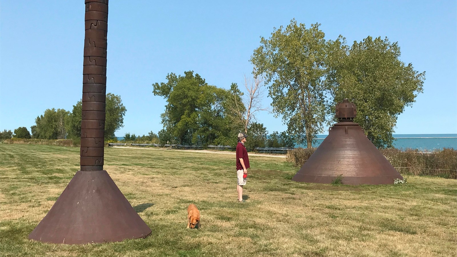 A man and his dog stand between two large industrial equipment on a grassy area.