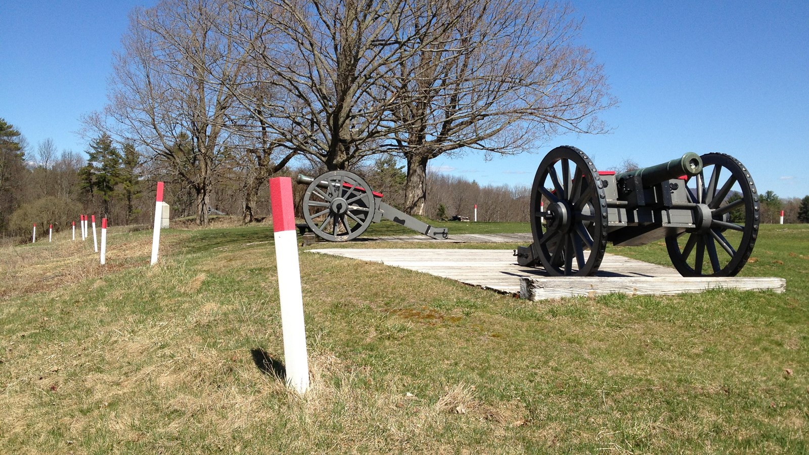 A cannon rests on a concrete pad next to a row of red topped, white marker posts. 