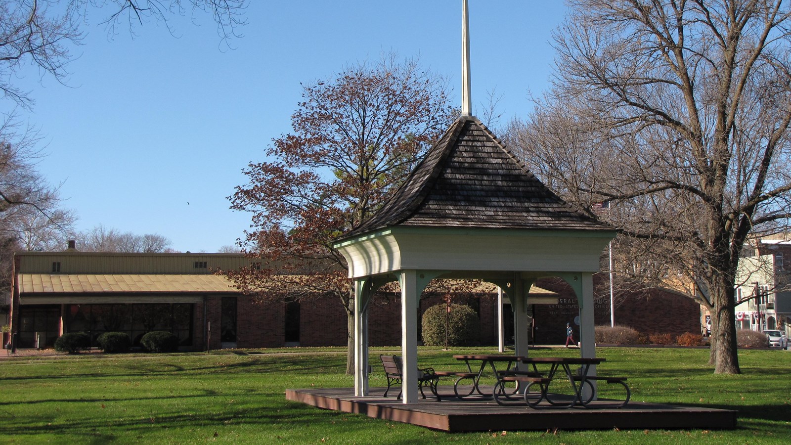 Trees shade a grassy park with a white gazebo in a historic downtown.