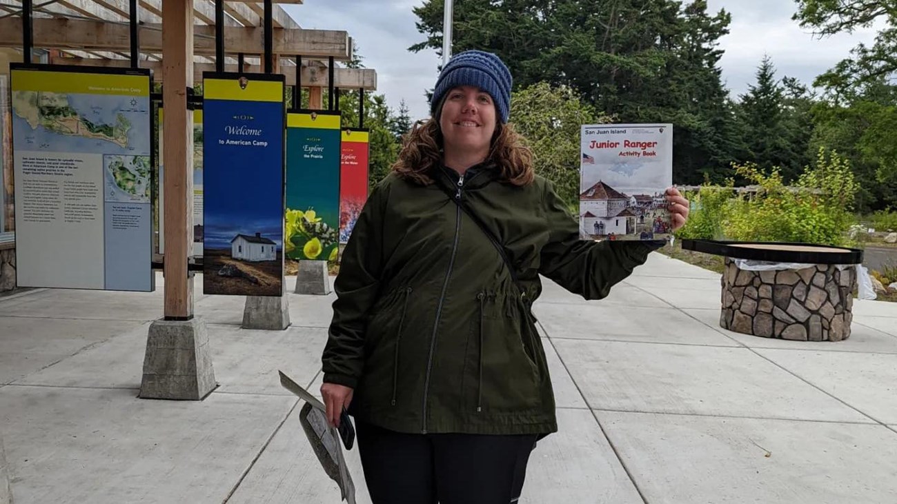 femme looking person in a coat and hat holds up a junior ranger booklet and is smiling