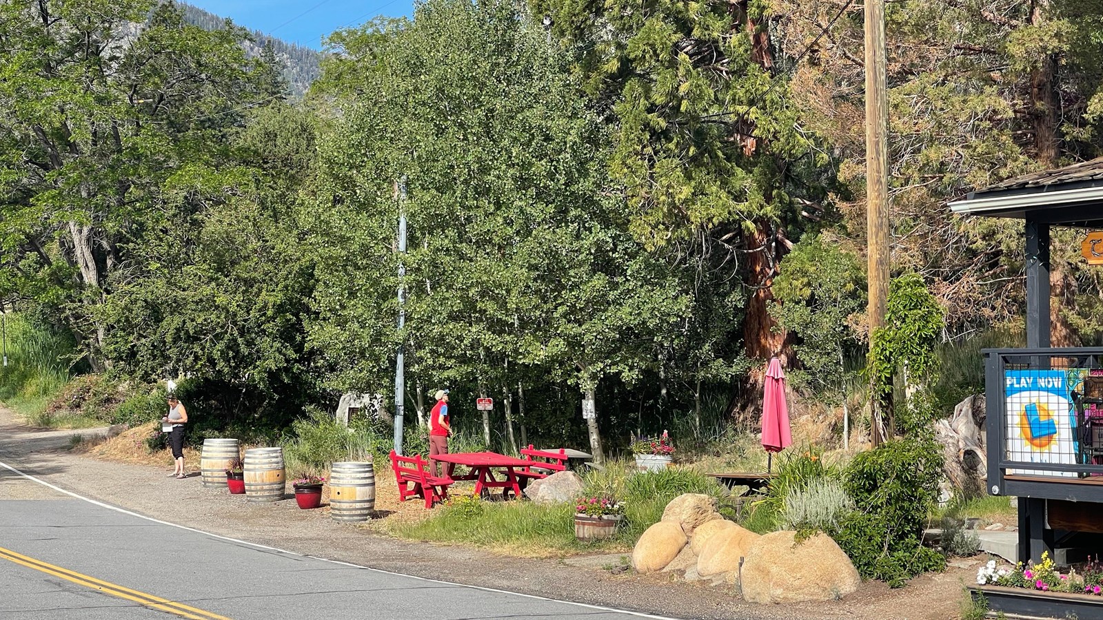 Two people stand near wood barrels in front of red picnic tables. A stone monument is among trees.