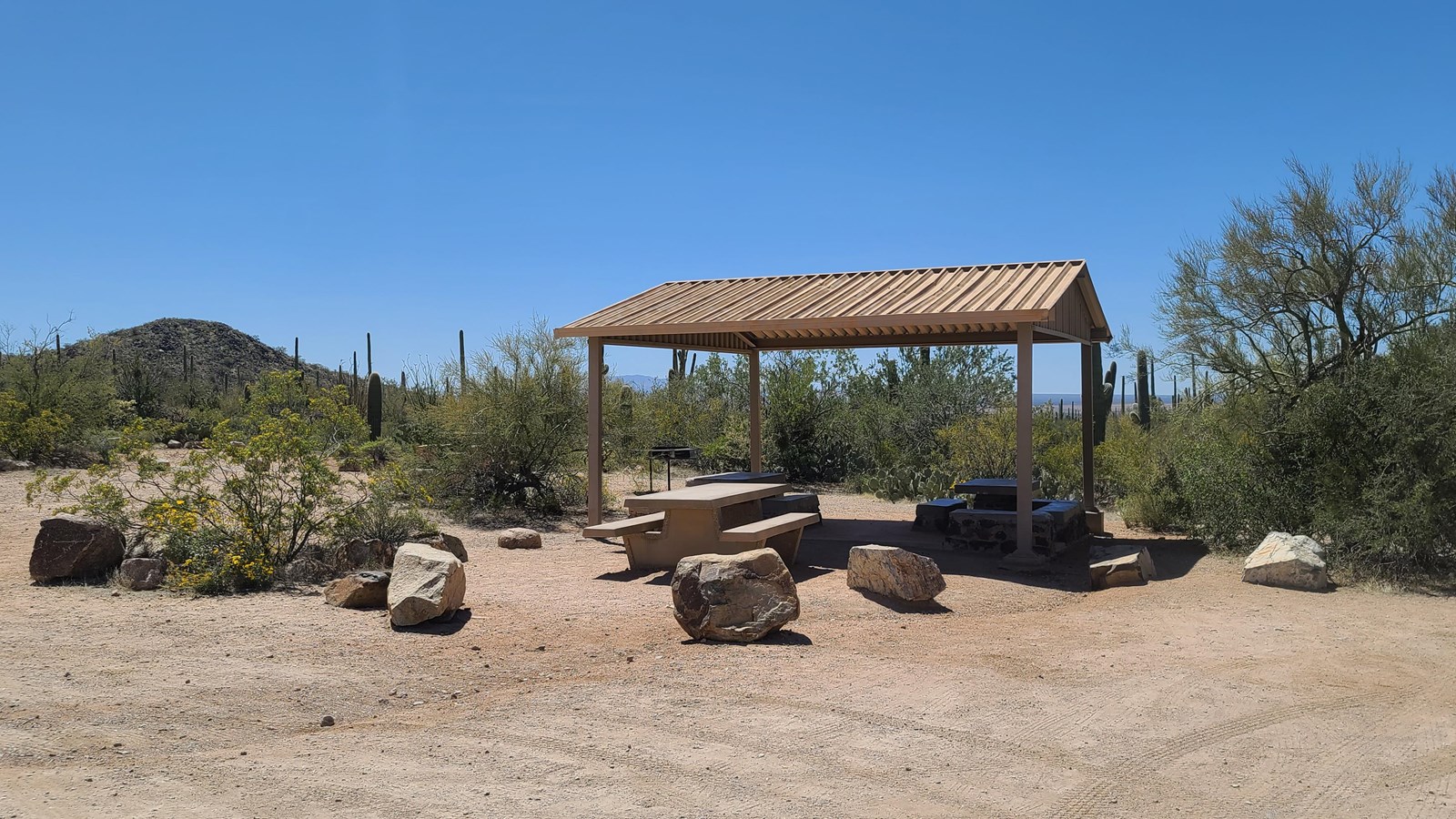 Three picnic tables and a shade structure surrounded by desert vegetation. 