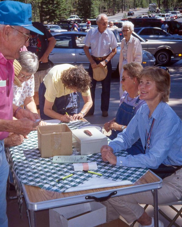 A seated woman smiles at an elderly man in a pink shirt at a table for a special event next to a par