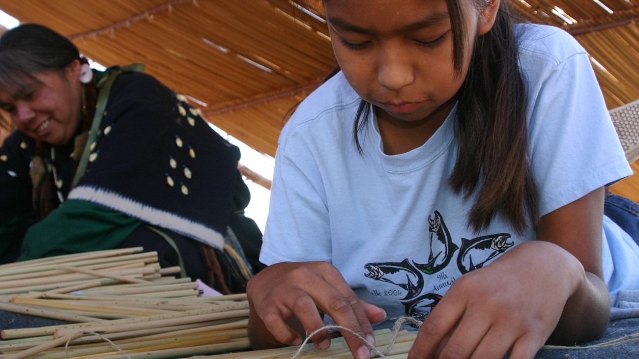 A color photo of a young girl leaning forward over a table and weaving rope around dried tule reeds.