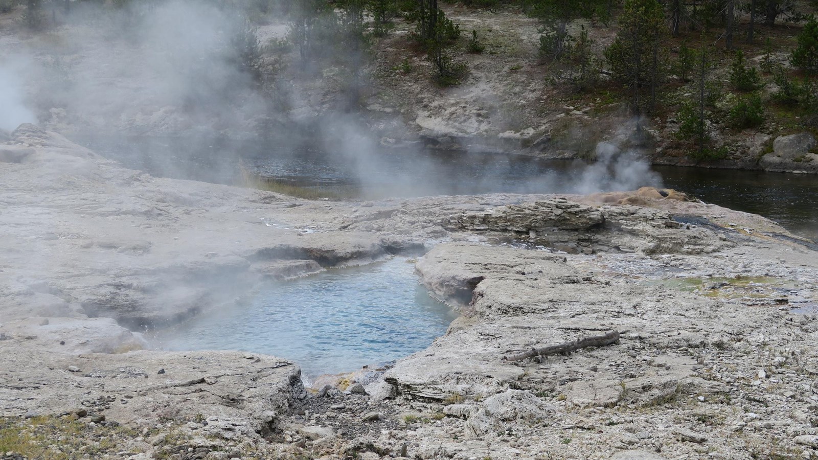 Steam rises from a hot spring in the sinter alongside a river bank.