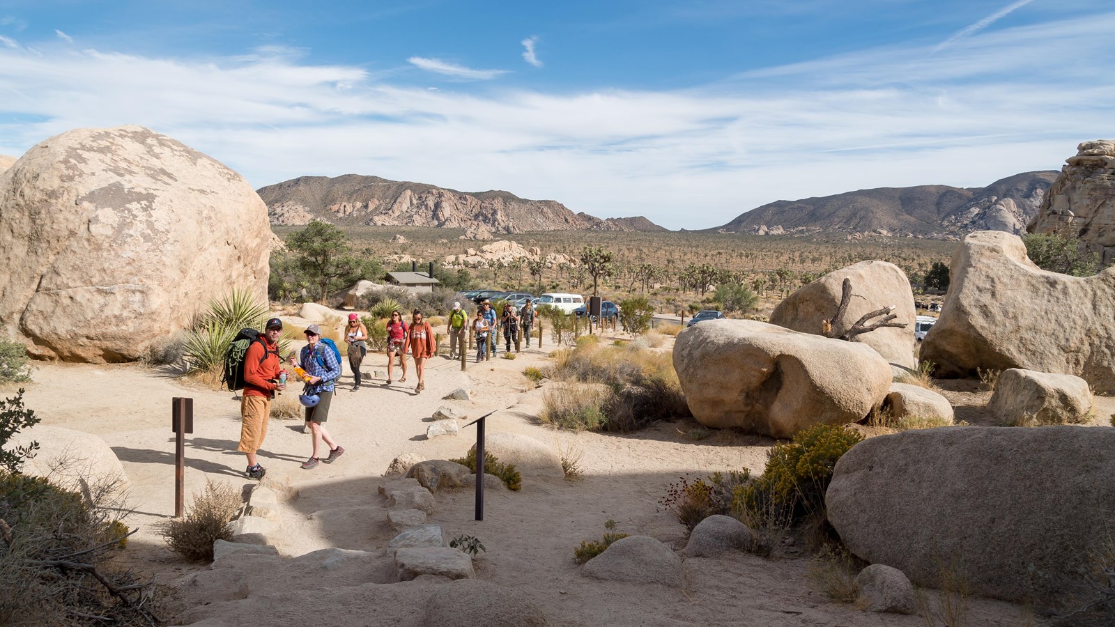 A group of hikers heading up a dirt trail between large rocks with mountains in the background.